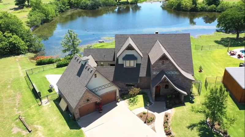 Aerial view of a large home with a shingle roof beside a lake